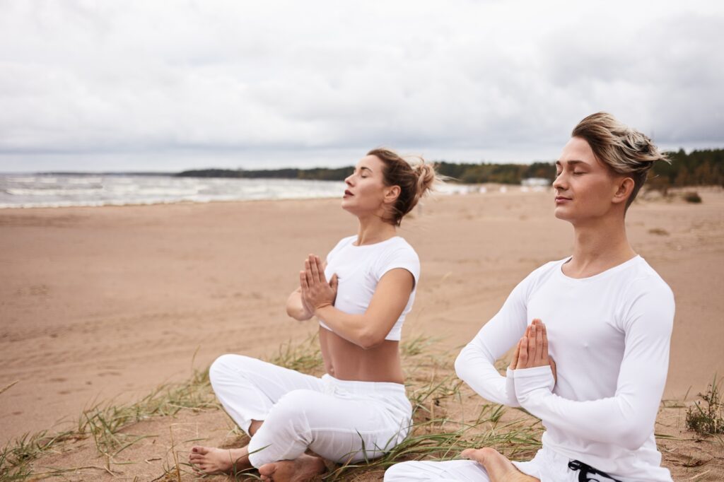 two atheltic european people man woman white sportswear closing eyes holding hands namste gesture sitting padmasana while meditating outdoors yoga retreat by ocean 1